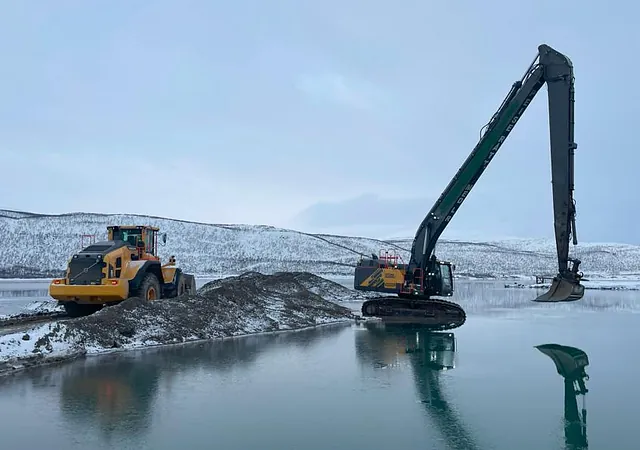 Gravemaskin med lang arm arbeider ved vannkant, ved siden av hjullaster i snødekt landskap.