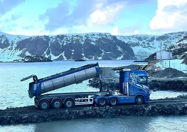 Tippbil med hevet lasteplan ved vannet, omgitt av snødekte fjell i kaldt landskap.