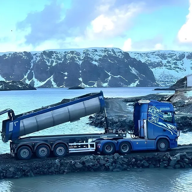 Tippbil med hevet lasteplan ved vannet, omgitt av snødekte fjell i kaldt landskap.