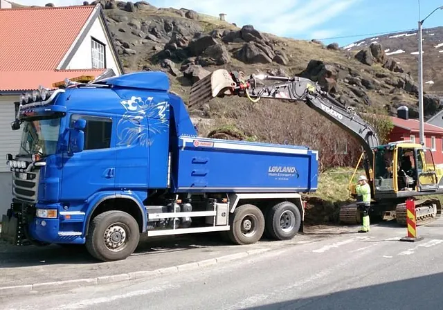 Gravemaskin laster masse på blå lastebil i boligområde med fjell og hus i bakgrunnen.